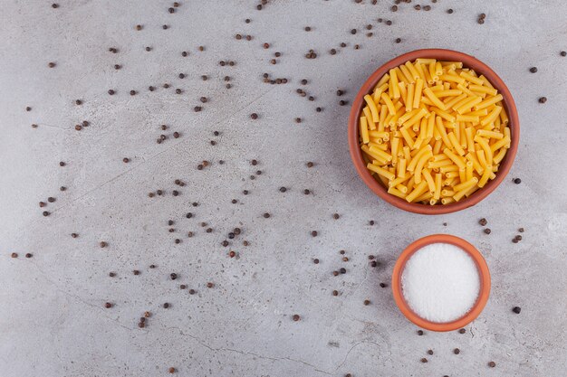 Dried penne Italian pasta in a round bowl with pepper corns on a stone table .