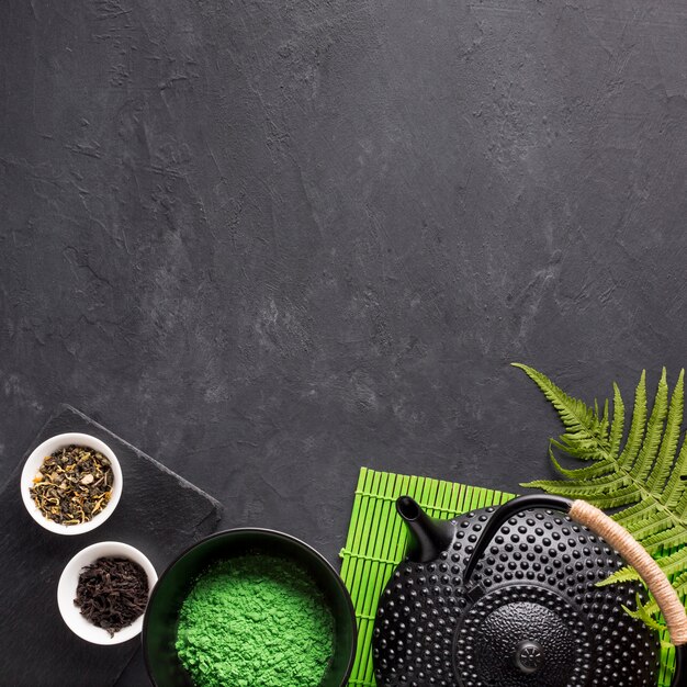 Dried tea herb and green matcha tea powder with teapot on black textured background