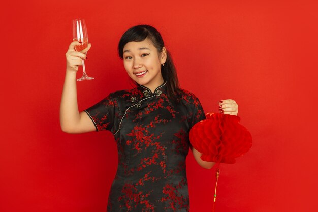 シャンパンを飲み、提灯を持っています。ハッピーチャイニーズニューイヤー2020。赤い背景の上のアジアの若い女の子の肖像画。伝統的な服を着た女性モデルは幸せそうに見えます。お祝い、感情。コピースペース。