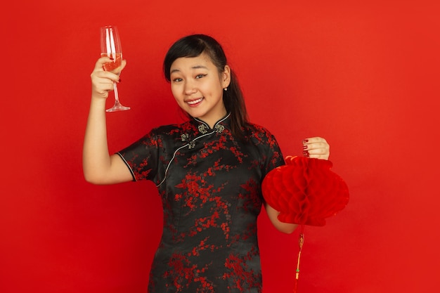 Drinking champagne and holding lantern. Happy Chinese New Year 2020. Asian young girl's portrait on red background. Female model in traditional clothes looks happy. Celebration, emotions. Copyspace.