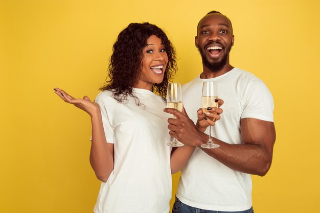 シャンパンを飲む。バレンタインデーのお祝い、黄色のスタジオの背景に分離された幸せなアフリカ系アメリカ人のカップル。人間の感情、顔の表情、愛、関係、ロマンチックな休日の概念。