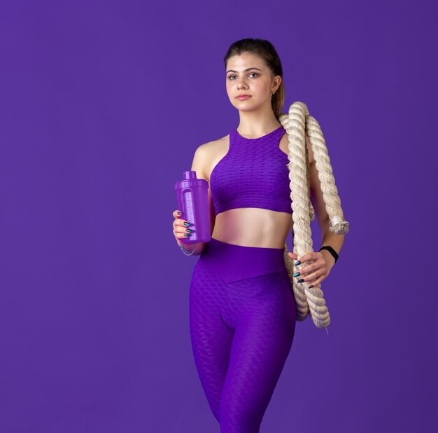 Drinking water with bottle. Beautiful young female athlete practicing , monochrome purple portrait. Sportive fit model with ropes. Body building, healthy lifestyle, beauty and action concept.