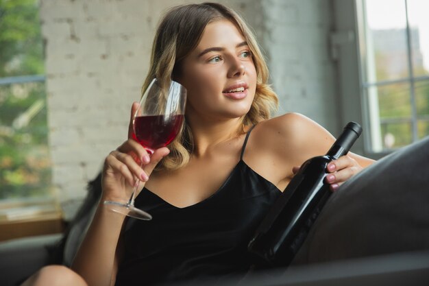 Drinking wine, looks cheerful. Portrait of pretty young girl in modern apartment in the morning.