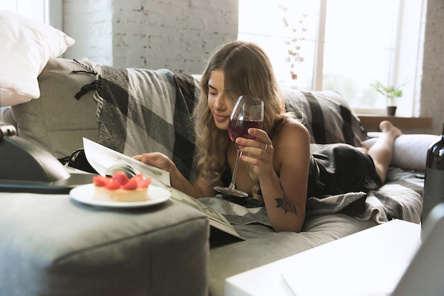 Free photo drinking wine, reading magazine. portrait of pretty young girl in modern apartment in the morning.