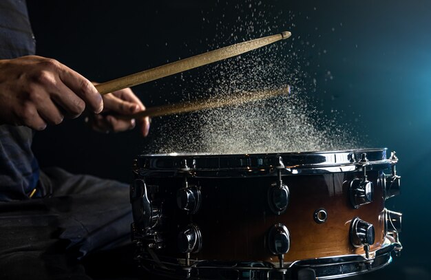 Drummer using drum sticks hitting snare drum with splashing water on black background under studio lighting close up.