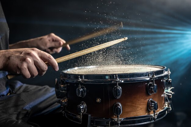 Drummer using drum sticks hitting snare drum with splashing water on black background under studio lighting close up.
