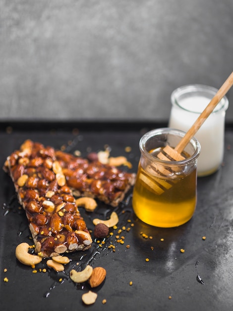 Dry fruit granola bars with honey and milk pots on textured black backdrop