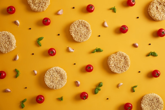 Dry instant noodles with tomatoes, garlic, basil and pepper for preparing fresh soup, yellow background. Preparing qucik lunch. Unhealthy eating and fast food concept. Ingredients for making dish