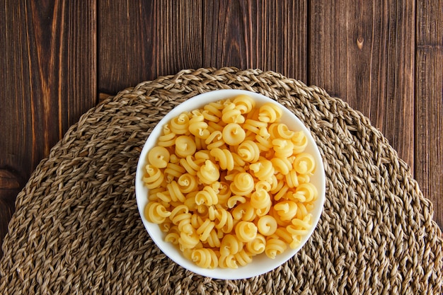 Free photo dry pasta in a bowl on wooden and wicker placemat background. flat lay.