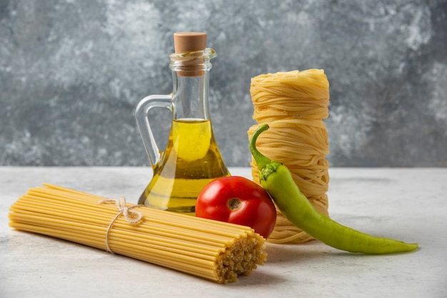 Dry pasta nests, spaghetti, bottle of olive oil and vegetables on white table. 