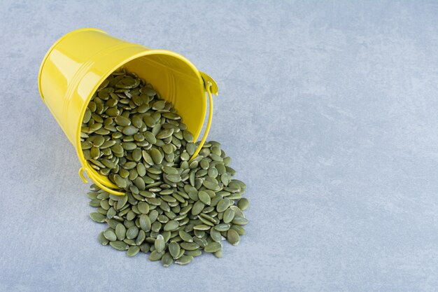 Free photo dry pepitas pouring out of a yellow bucket on marble background.