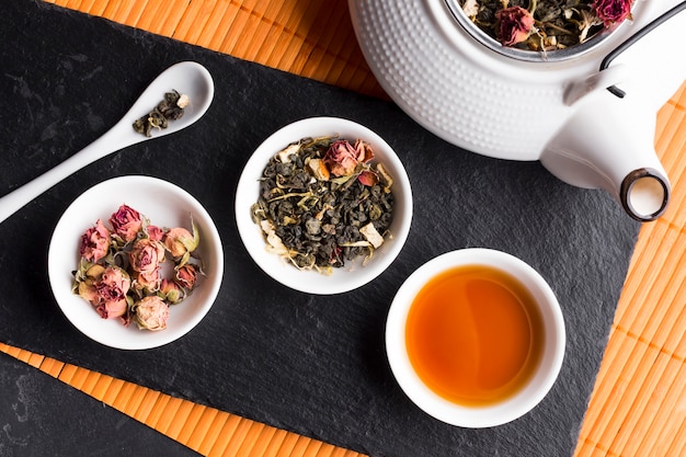 Free photo dry roses and tea herb with teapot on black slate over placemat