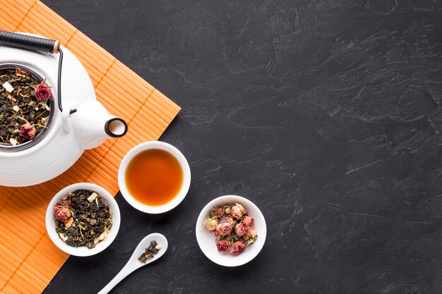 Dry roses and tea herb with teapot on orange placemat over black stone backdrop