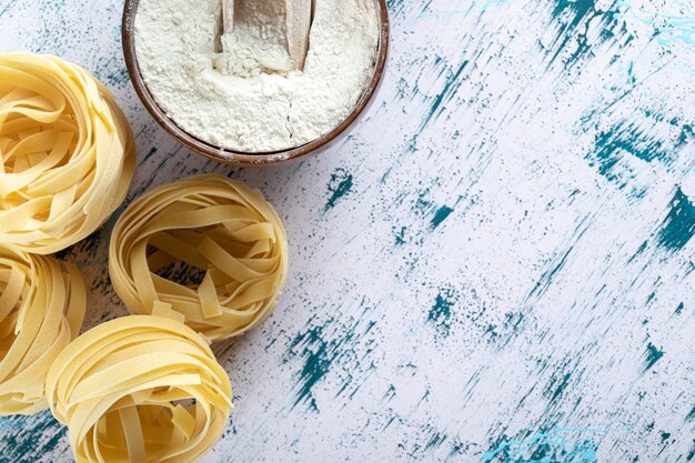 Dry tagliatelle nests and bowl of flour on colorful surface.