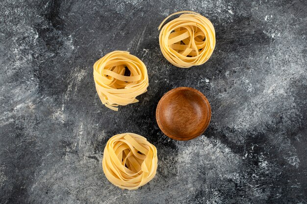 Dry tagliatelle nests and wooden bowl on marble surface