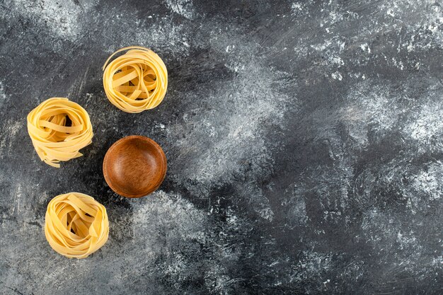 Dry tagliatelle nests and wooden bowl on marble surface