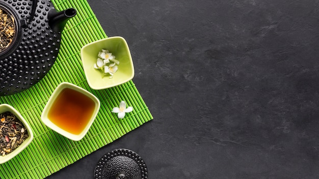Бесплатное фото Сухая чайная трава и травяной чай с белым цветком жасмина на зеленом столовом приборе на черном фоне