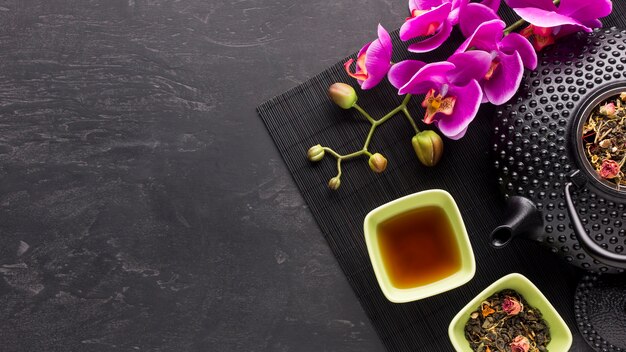 Dry tea herb and pink orchid flower with teapot on black surface