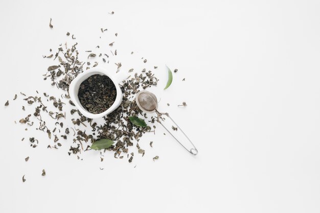 Dry tea leaves with coffee leaves and tea strainer on white backdrop
