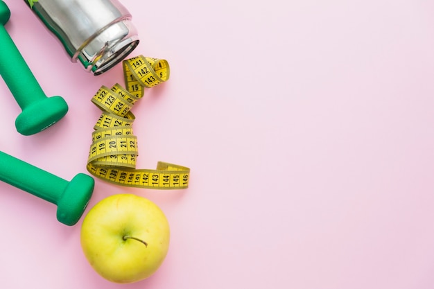 Dumbbells; water bottle; measuring tape and apple on pink background