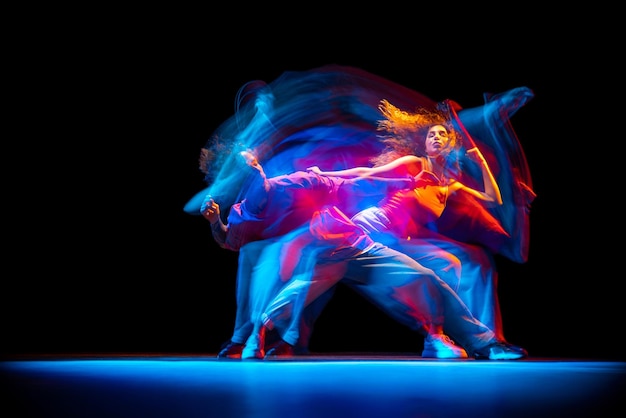 Dynamic portrait of young man and woman dancing hiphop isolated over black background with mixed lights effect