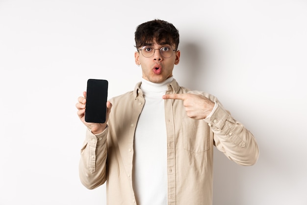 E-commerce concept. Portrait of young man pointing at mobile phone screen, showing advertisement online, standing on white background
