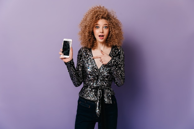 Free photo eager woman with blue eyes and curly hair dressed in glittering disco blouse points to smartphone.