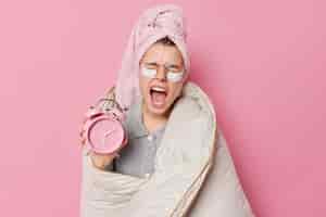 Бесплатное фото Концепция раннего пробуждения. сонная молодая европейка зевает и широко открывает рот, держит будильник, завернутый в одеяло, носит банное полотенце на голове после принятия душа на розовом фоне