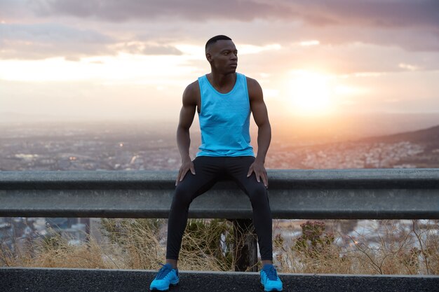 Early morning sport concept. Thoughtful black ethnic man sits at traffic sign, poses against magnificent sunrise view, enjoys calm atmosphere, wears blue vest, black trousers and sportshoes.