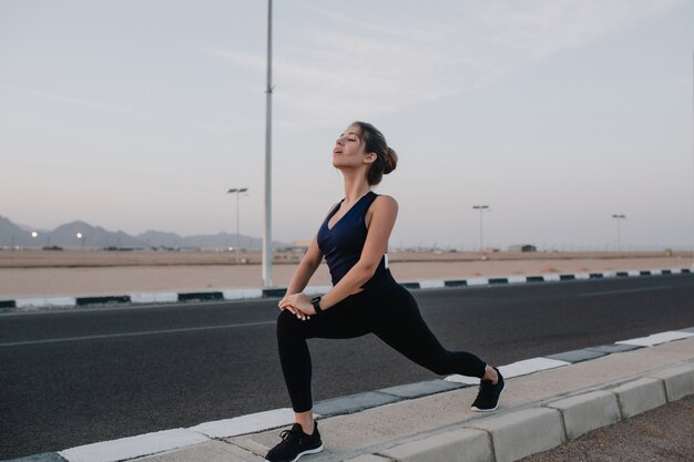 Early morning time for training of young pretty woman stretching on road of tropical country. Summer period, enjoying sport, relaxing.