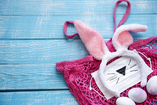 Easter composition with pink string bag, decorative Easter bunny ears, medical mask and eggs on a wooden surface