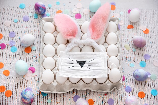 Easter composition with a tray of eggs, a medical mask with a painted Easter bunny face and ears among confetti close up