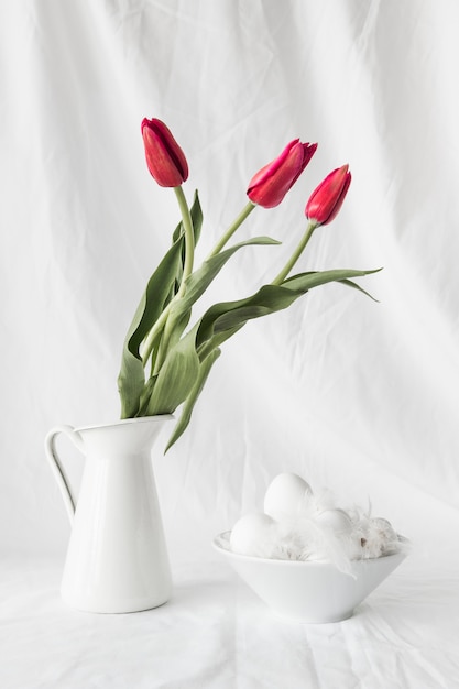 Easter eggs and quills in bowl near bunch of flowers in vase