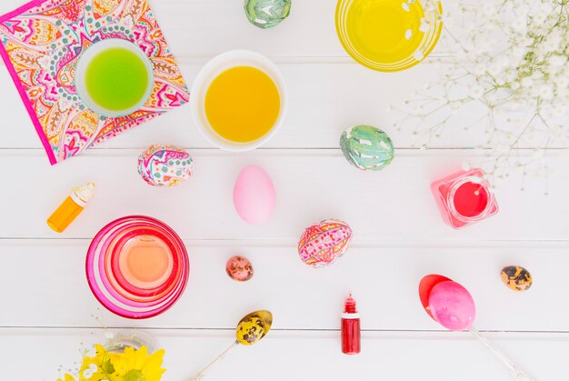 Easter eggs between spoons near napkin, flower twig and cups with dye liquid