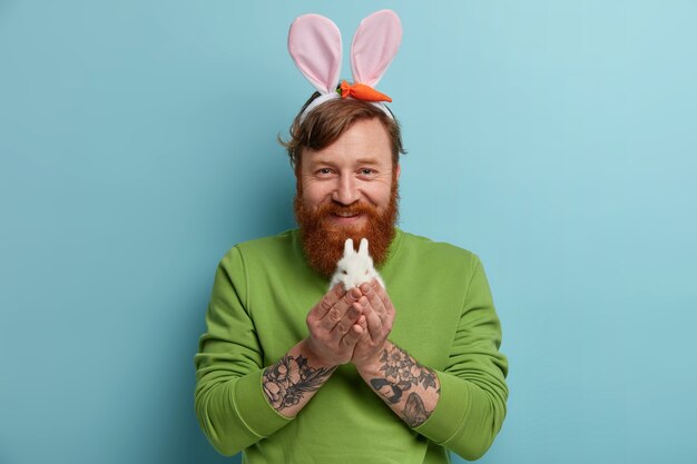 Easter holiday concept. Bearded ginger man with tattooed arms holds tiny white fluffy rabbit, wears bunny ears, green jumper, has happy expression, isolated over blue wall. Easter symbol