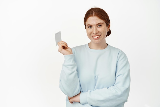 Easy payment. Smiling ginger girl look confident, holding credit discount card and looking happy, going on shopping, paying contactless, white background.