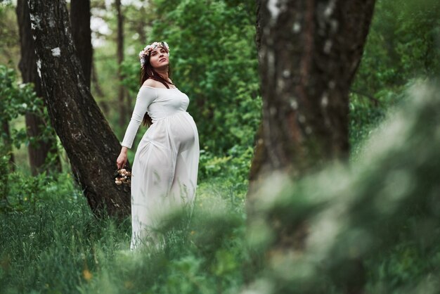 Easy wind going through the woods. Beautiful pregnant woman in dress have a walk outdoors. Positive brunette