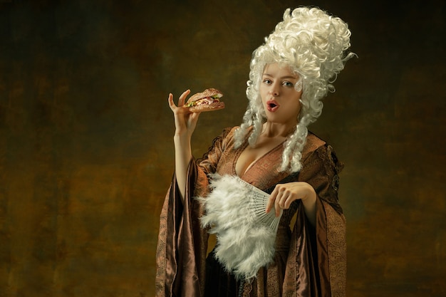 Eating burger delighted. Portrait of medieval young woman in brown vintage clothing on dark background. Female model as a duchess, royal person. Concept of comparison of eras, modern, fashion, beauty.