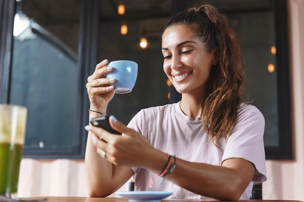 Eating out and lifestyle concept Happy caucasian woman with tropical tan drinking coffee and checking messages on mobile phone having breakfast on terrace and smiling