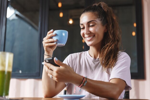 Eating out and lifestyle concept Happy caucasian woman with tropical tan drinking coffee and checking messages on mobile phone having breakfast on terrace and smiling
