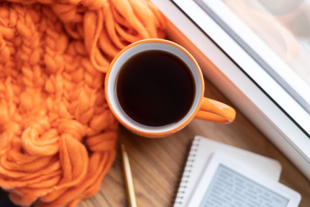 An ebook and a pen with a notepad and a mug of coffee tea on an orange knitted sweater scarf plaid on the windowsill