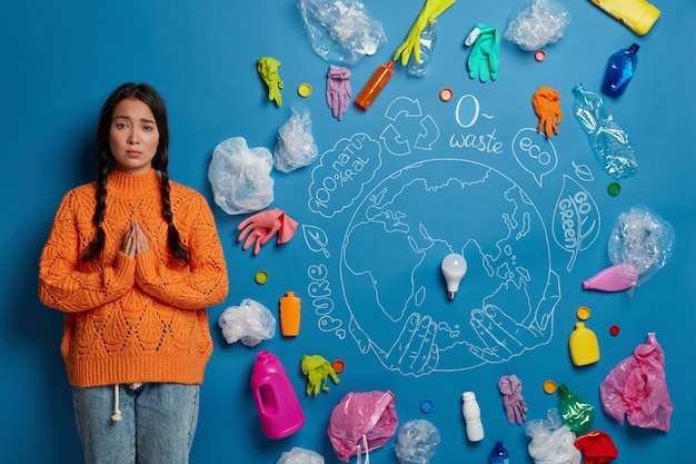 Ecology and land conservation concept. Sad Asian woman stands in praying pose, surrounded with plastic waste, begs for help in cleaning Earth, dressed casually