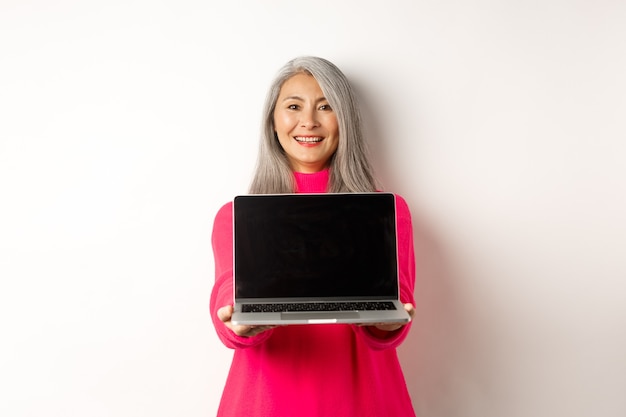 空白のノートパソコンの画面を表示し、幸せなデモを探しているアジアの年配の女性を笑顔のeコマースの概念...