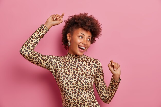 Ecsatitic overjoyed dark skinned woman with curly hair, keeps hands raised, dances carefree, celebrates victory or success, wears leopard turtleneck