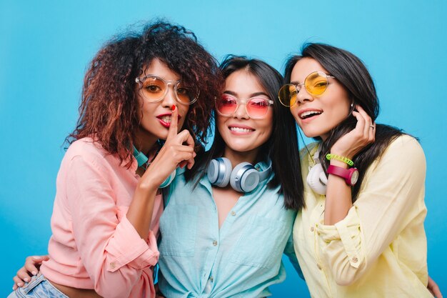 Ecstatic asian girl in blue cotton shirt embracing friends. Indoor portrait of catching african lady with curly hair having fun with female colleagues.