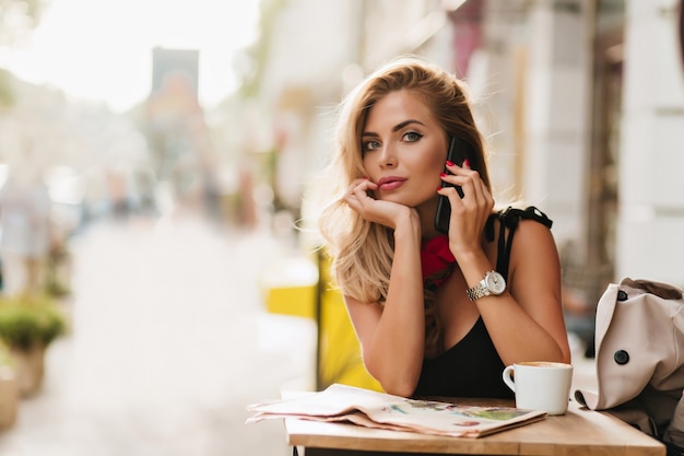 Ecstatic blonde woman talking on phone, propping face with hand after drinking coffee