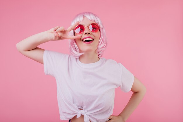 Ragazza caucasica estatica in maglietta bianca alla moda in posa con segno di pace e ridendo. foto dell'interno di una donna europea sognante in peruke lucido e occhiali da sole