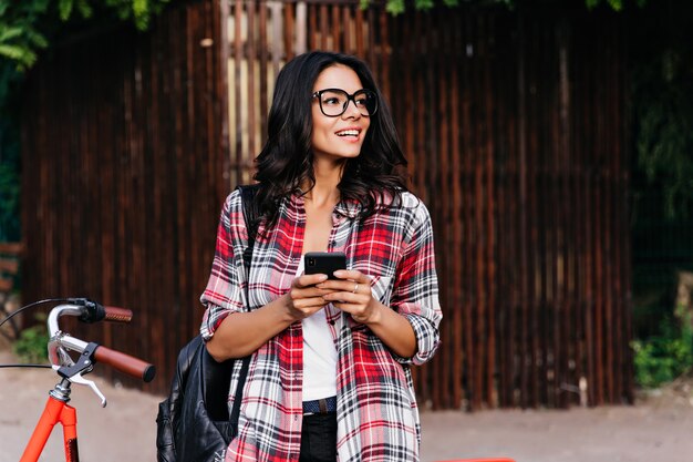 Восторженная кудрявая девушка в красной клетчатой рубашке, стоя перед велосипедом. Открытый портрет добродушной латинской дамы с телефоном, оглядывающей улицу.