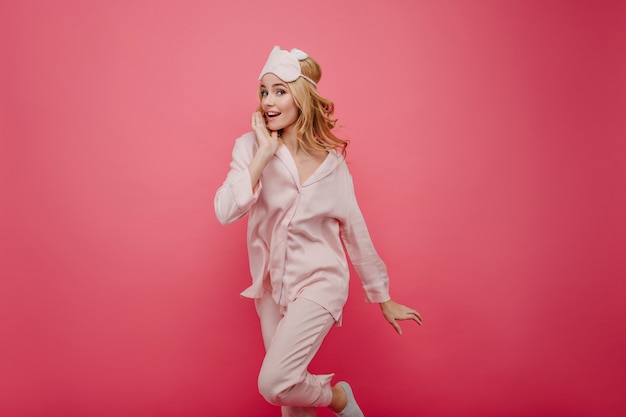 Ecstatic curly girl in silk pajama jumping on bright wall. Emotional lady in sleepmask having fun  with pink interior.