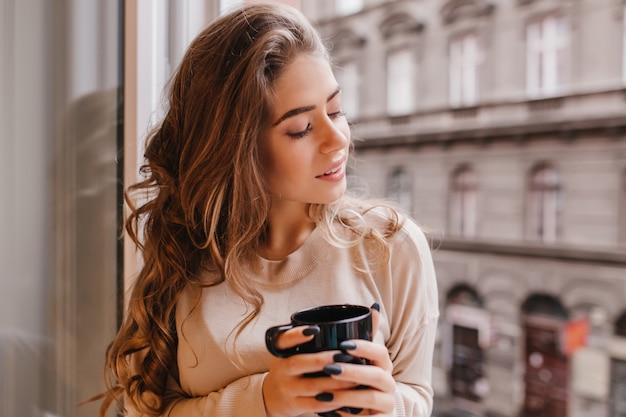 Ecstatic curly woman sitting with eyes closed near big window